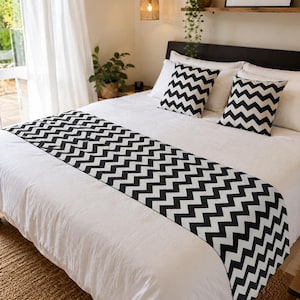 Könnte beinhalten: Ein Bettläufer und zwei quadratische Dekokissen mit einem schwarz-weißen Zickzack-Chevron-Muster. Die Textilien liegen auf einer weißen Tagesdecke und bilden ein kontrastreiches, geometrisches Designelement für das Bett-Set.