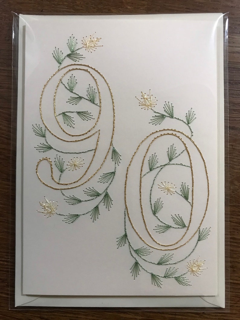 K&ouml;nnte beinhalten: Eine wei&szlig;e Gru&szlig;karte mit der Zahl 90, gestickt mit goldenem Faden. Die Zahlen sind mit gr&uuml;nen und goldenen floralen Akzenten verziert. Die Karte ist in einer transparenten Plastikh&uuml;lle verpackt.