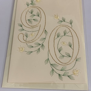 K&ouml;nnte beinhalten: Cremefarbene Gru&szlig;karte mit der Zahl 90, gestickt mit goldenem Faden, verziert mit gr&uuml;nen und goldenen floralen Akzenten. Die Karte befindet sich in einer transparenten Plastikh&uuml;lle, passend f&uuml;r einen runden Geburtstag.