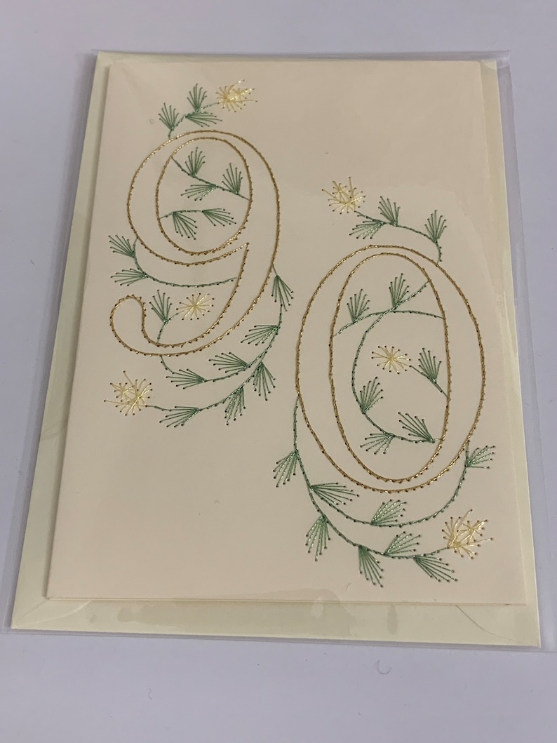 K&ouml;nnte beinhalten: Cremefarbene Gru&szlig;karte mit der Zahl 90, gestickt mit goldenem Faden, verziert mit gr&uuml;nen und goldenen floralen Akzenten. Die Karte befindet sich in einer transparenten Plastikh&uuml;lle, passend f&uuml;r einen runden Geburtstag.
