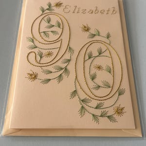 K&ouml;nnte beinhalten: Handgefertigte Gru&szlig;karte mit dem Namen "Elizabeth" in hellbraunem Faden gestickt. Die Zahl "90" ist mit goldenem Faden gestickt, verziert mit gr&uuml;nen Bl&auml;ttern und kleinen gelben Bl&uuml;ten.
