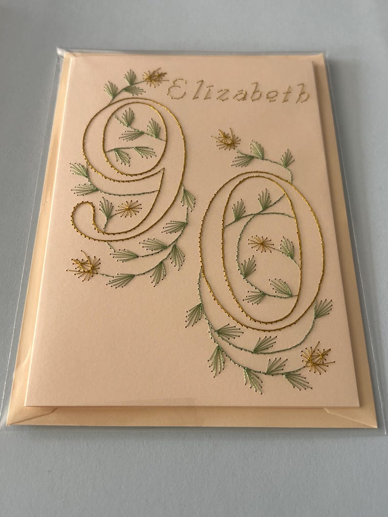 K&ouml;nnte beinhalten: Handgefertigte Gru&szlig;karte mit dem Namen "Elizabeth" in hellbraunem Faden gestickt. Die Zahl "90" ist mit goldenem Faden gestickt, verziert mit gr&uuml;nen Bl&auml;ttern und kleinen gelben Bl&uuml;ten.
