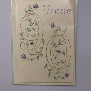 K&ouml;nnte beinhalten: Handgefertigte Gru&szlig;karte mit dem Namen "Irene" und der Zahl "90", gestickt mit gold-, gr&uuml;n- und lilafarbenem Faden. Die cremefarbene Karte hat florale Akzente und ist in einer transparenten Plastikh&uuml;lle.