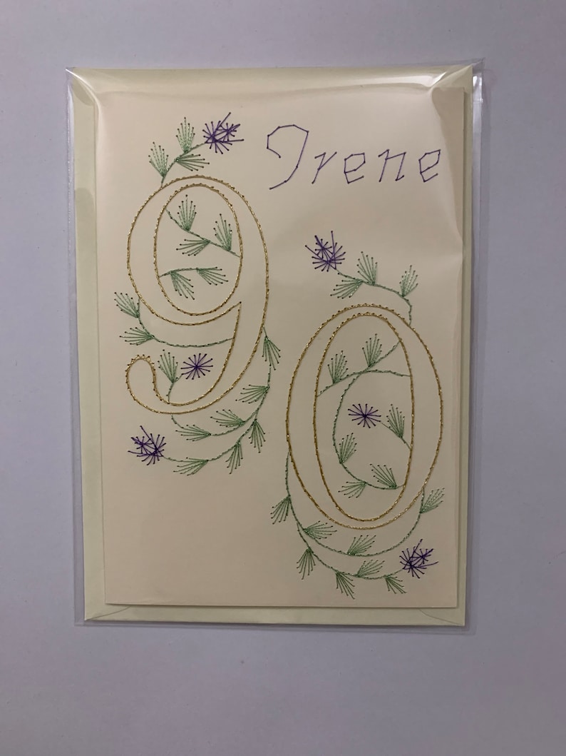 K&ouml;nnte beinhalten: Handgefertigte Gru&szlig;karte mit dem Namen "Irene" und der Zahl "90", gestickt mit gold-, gr&uuml;n- und lilafarbenem Faden. Die cremefarbene Karte hat florale Akzente und ist in einer transparenten Plastikh&uuml;lle.
