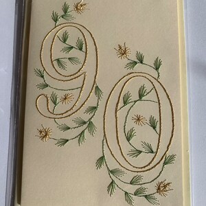 K&ouml;nnte beinhalten: Cremefarbene Gru&szlig;karte mit der Zahl "90" in goldener Schrift, verziert mit gr&uuml;ner und goldener Blumenstickerei. Die Karte ist in einer transparenten Plastikh&uuml;lle eingeschlossen.
