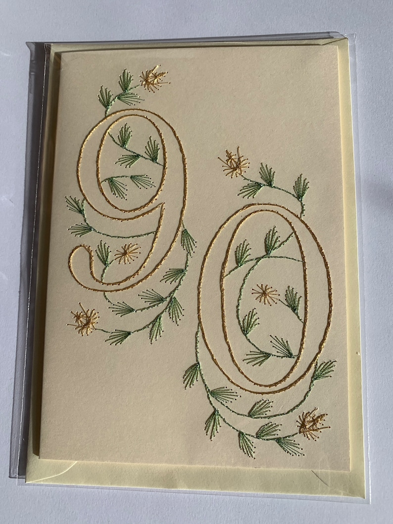 K&ouml;nnte beinhalten: Cremefarbene Gru&szlig;karte mit der Zahl "90" in goldener Schrift, verziert mit gr&uuml;ner und goldener Blumenstickerei. Die Karte ist in einer transparenten Plastikh&uuml;lle eingeschlossen.