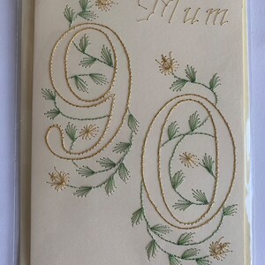 K&ouml;nnte beinhalten: Cremefarbene Gru&szlig;karte mit dem Wort "Mum" in Goldfaden gestickt. Die Zahlen "90" sind ebenfalls mit Goldfaden gestickt, mit gr&uuml;nen und goldenen floralen Akzenten. Die Karte befindet sich in einer transparenten Plastikh&uuml;lle.