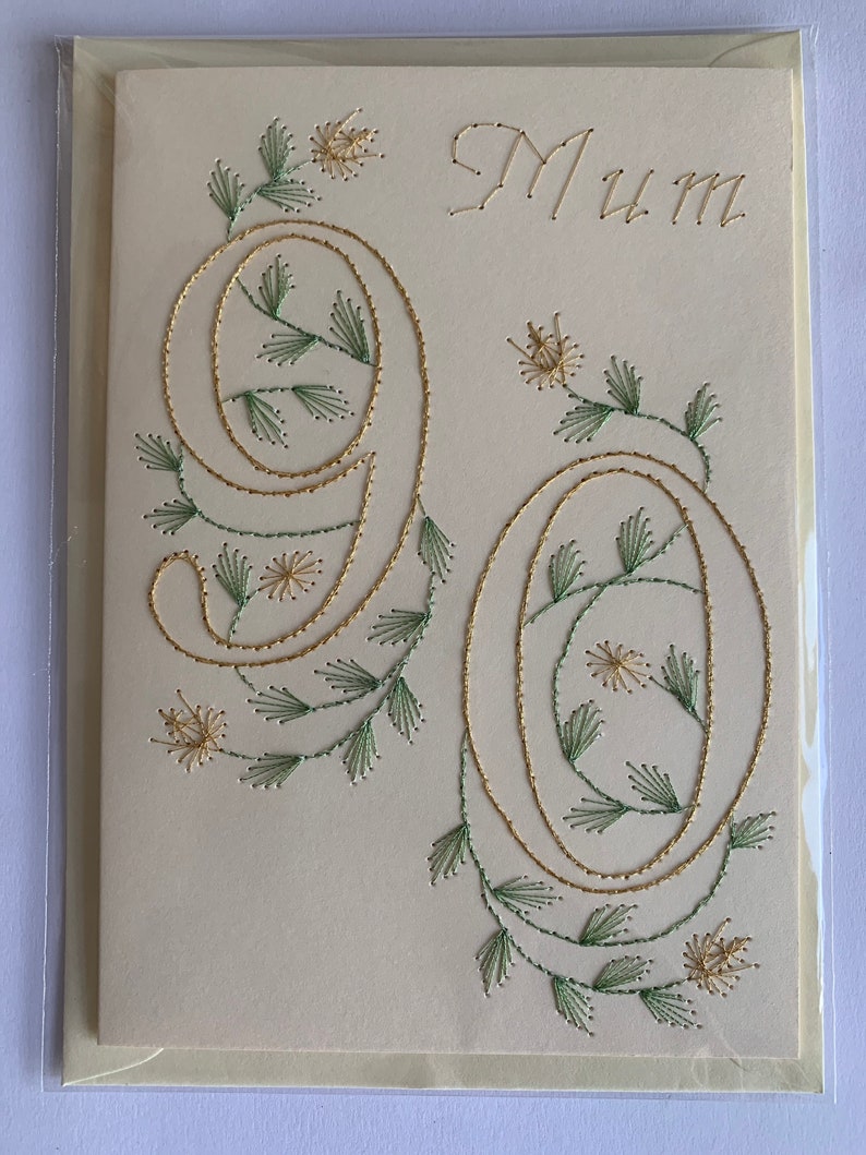 K&ouml;nnte beinhalten: Cremefarbene Gru&szlig;karte mit dem Wort "Mum" in Goldfaden gestickt. Die Zahlen "90" sind ebenfalls mit Goldfaden gestickt, mit gr&uuml;nen und goldenen floralen Akzenten. Die Karte befindet sich in einer transparenten Plastikh&uuml;lle.