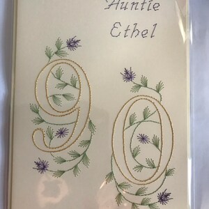 K&ouml;nnte beinhalten: Cremefarbene Gru&szlig;karte mit den Worten "Auntie Ethel" in lila Schrift. Die Zahl "90" ist mit goldfarbenem Faden bestickt, mit gr&uuml;nen Bl&auml;ttern und lila Blumen. Die Karte befindet sich in einer transparenten Plastikh&uuml;lle.