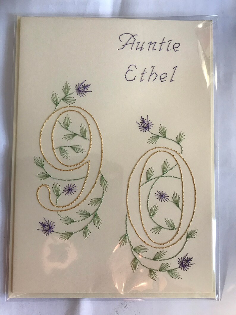 K&ouml;nnte beinhalten: Cremefarbene Gru&szlig;karte mit den Worten "Auntie Ethel" in lila Schrift. Die Zahl "90" ist mit goldfarbenem Faden bestickt, mit gr&uuml;nen Bl&auml;ttern und lila Blumen. Die Karte befindet sich in einer transparenten Plastikh&uuml;lle.