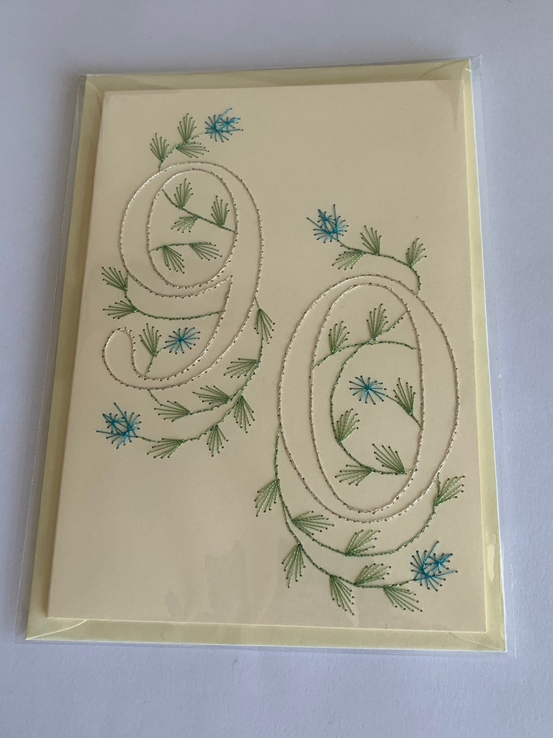 K&ouml;nnte beinhalten: Cremefarbene Gru&szlig;karte mit den gestickten Zahlen "90". Die Zahlen sind mit silbernem Faden umrandet und mit gr&uuml;nen und blauen floralen Akzenten verziert. Die Karte befindet sich in einer transparenten Plastikh&uuml;lle.