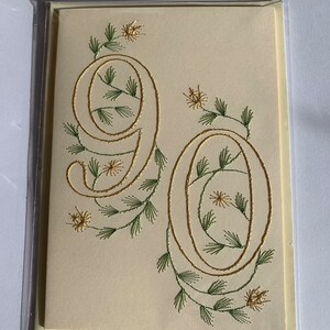 K&ouml;nnte beinhalten: Cremefarbene Gru&szlig;karte mit der Zahl 90, die mit goldenem Faden gestickt ist. Die Zahlen sind mit gr&uuml;nen und goldenen floralen Akzenten verziert, was ein elegantes Design ergibt. Die Karte ist in einer transparenten Plastikh&uuml;lle eingeschlossen.