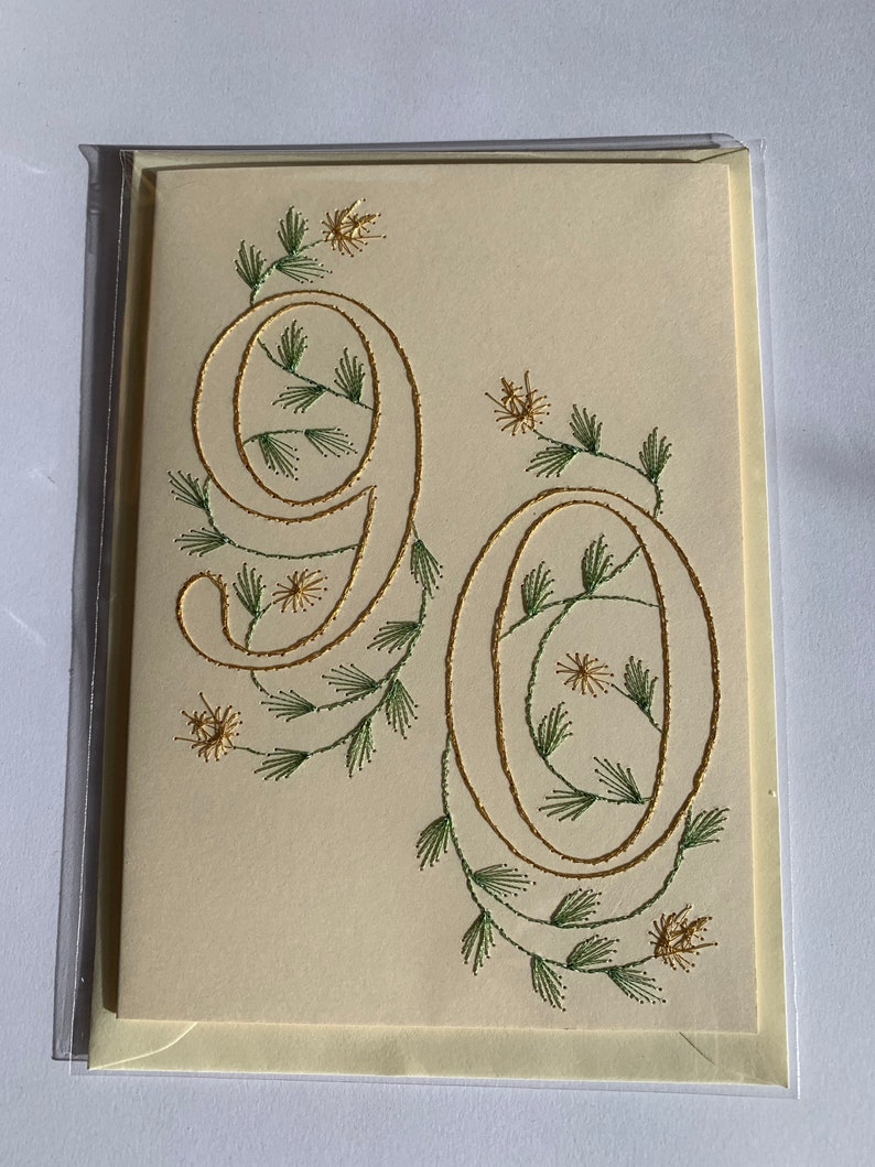 K&ouml;nnte beinhalten: Cremefarbene Gru&szlig;karte mit der Zahl 90, die mit goldenem Faden gestickt ist. Die Zahlen sind mit gr&uuml;nen und goldenen floralen Akzenten verziert, was ein elegantes Design ergibt. Die Karte ist in einer transparenten Plastikh&uuml;lle eingeschlossen.