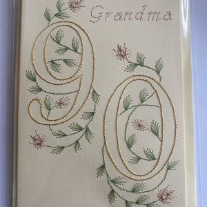 K&ouml;nnte beinhalten: Cremefarbene Gru&szlig;karte mit dem Wort "Oma" in braunem Faden gestickt. Die Zahl "90" ist mit goldenem Faden bestickt, verziert mit gr&uuml;nen und rosa floralen Akzenten. Die Karte befindet sich in einer transparenten Plastikh&uuml;lle.