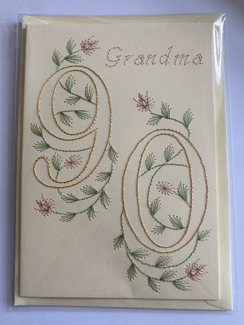 K&ouml;nnte beinhalten: Cremefarbene Gru&szlig;karte mit dem Wort "Oma" in braunem Faden gestickt. Die Zahl "90" ist mit goldenem Faden bestickt, verziert mit gr&uuml;nen und rosa floralen Akzenten. Die Karte befindet sich in einer transparenten Plastikh&uuml;lle.