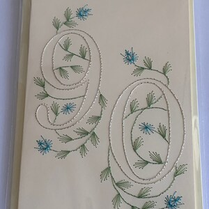 K&ouml;nnte beinhalten: Cremefarbene Gru&szlig;karte mit der gestickten Zahl 90. Die Zahlen sind mit silbernem Faden umrandet und mit gr&uuml;nen Bl&auml;ttern und blauen Blumen verziert. Die Karte befindet sich in einer transparenten Plastikh&uuml;lle.