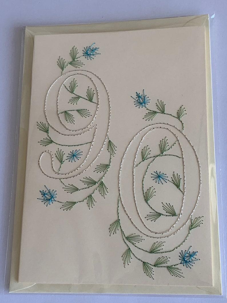 K&ouml;nnte beinhalten: Cremefarbene Gru&szlig;karte mit der gestickten Zahl 90. Die Zahlen sind mit silbernem Faden umrandet und mit gr&uuml;nen Bl&auml;ttern und blauen Blumen verziert. Die Karte befindet sich in einer transparenten Plastikh&uuml;lle.