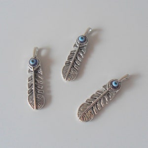 Puede incluir: Tres abalorios de plumas plateadas con detalles de ojo azul maligno. Las plumas están detalladas con líneas grabadas y tienen un bucle en la parte superior para sujetarlas a joyas.