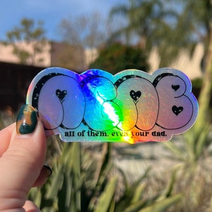 May include: A holographic sticker with the word "OG" in bubble letters and hearts. The sticker has the text "all of them. even your dad." below the word "OG".