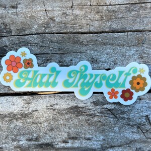 Puede incluir: Una pegatina blanca con el texto "Hair Thyself" en una fuente retro, con un fondo azul claro y un contorno amarillo. La pegatina está decorada con flores naranjas, amarillas y marrones.