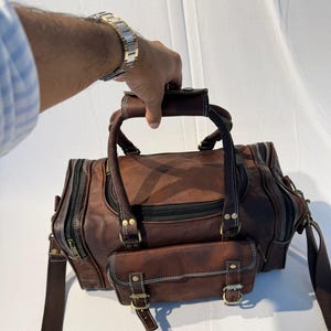 Puede incluir: Una bolsa de lona de cuero marrón con múltiples compartimentos y cremalleras. La bolsa tiene un asa superior y una correa para el hombro. La bolsa tiene un bolsillo frontal con hebillas y costuras blancas. La bolsa es sostenida por una persona.
