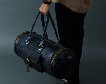 Può includere: Una borsa da viaggio in pelle blu scuro con una tasca frontale e dettagli dorati. La borsa ha una forma cilindrica ed è tenuta da una persona.
