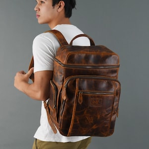 Może przedstawiać: Brązowy skórzany plecak z wieloma kieszeniami i przetartym wyglądem. Plecak ma zamek błyskawiczny i górny uchwyt.