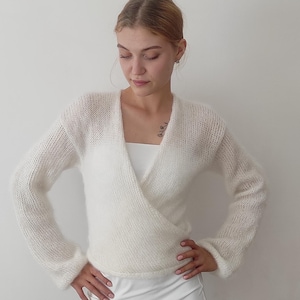 May include: A white, long-sleeved wrap sweater with a V-neck. The sweater is made of a soft, fuzzy material. The model is wearing a white top and a white skirt.