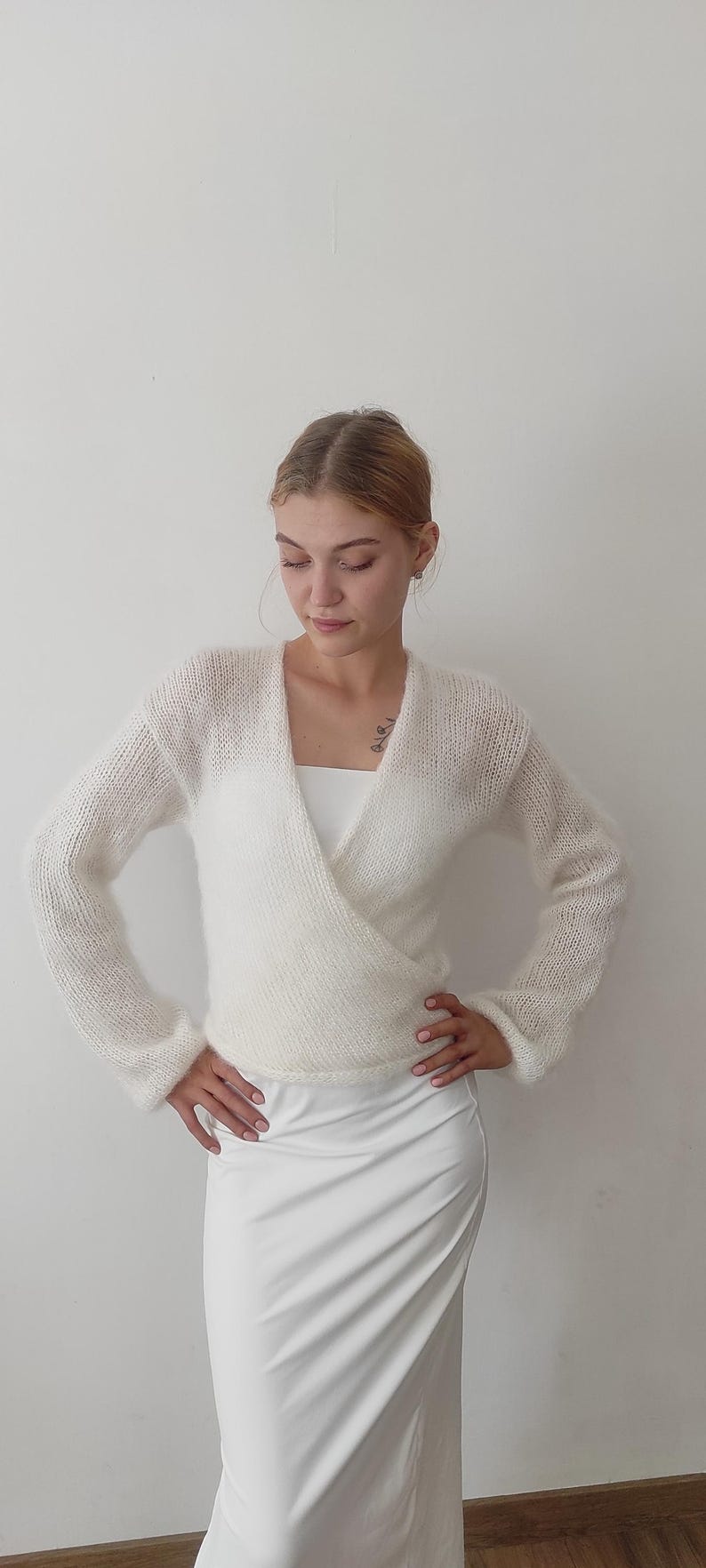 May include: A white, long-sleeved wrap sweater with a V-neck. The sweater is made of a soft, fuzzy material. The model is wearing a white top and a white skirt.