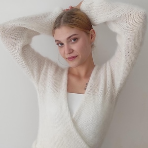 May include: A white, fluffy, wrap-style sweater with long sleeves. The sweater is paired with a white top and a white skirt. The model has their arms raised above their head.