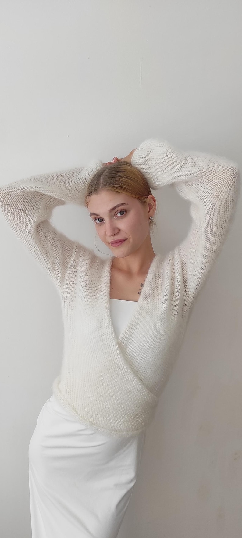 May include: A white, fluffy, wrap-style sweater with long sleeves. The sweater is paired with a white top and a white skirt. The model has their arms raised above their head.