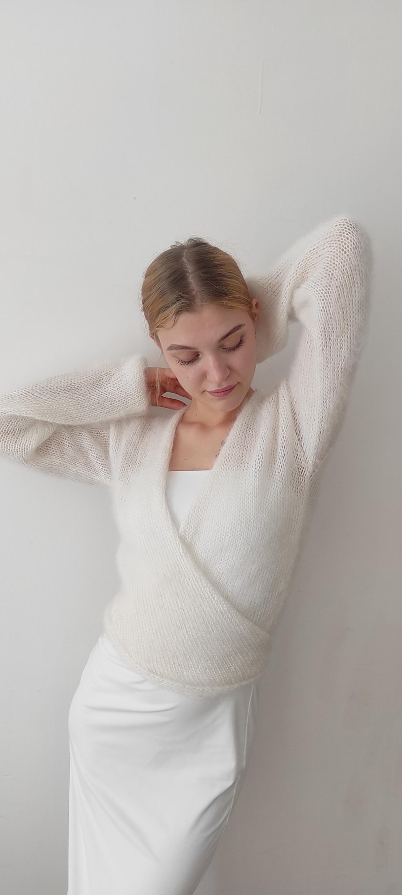 May include: A white, open-front wrap sweater with long sleeves. The sweater is made of a soft, fuzzy material. The model is wearing a white top and a white skirt. The background is a plain white wall.