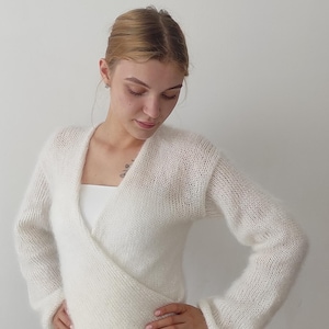 May include: A white, long-sleeved wrap sweater with a V-neck. The sweater is made of a soft, fuzzy material. The model is wearing a white dress underneath. The background is a plain white wall.