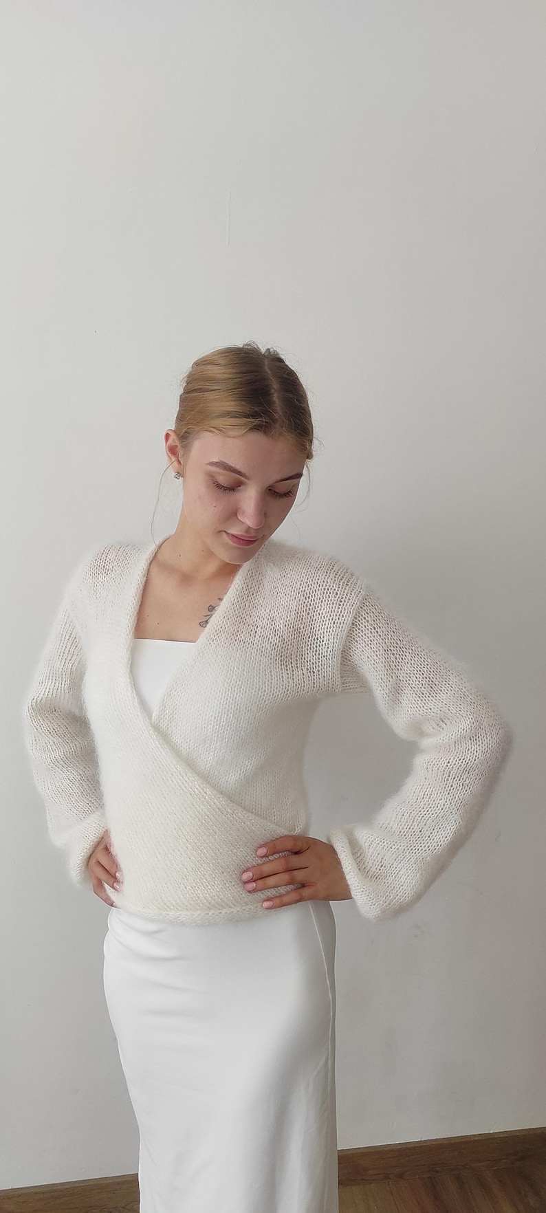 May include: A white, long-sleeved wrap sweater with a V-neck. The sweater is made of a soft, fuzzy material. The model is wearing a white dress underneath. The background is a plain white wall.