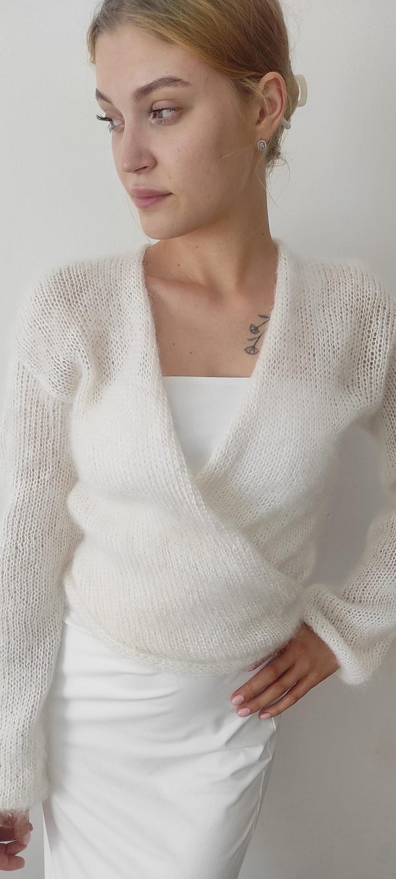 May include: A white, sheer, wrap-style sweater with long sleeves. The sweater is made of a soft, textured knit material. The model is wearing a white top underneath. The overall look is elegant and delicate.
