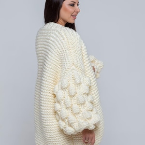 May include: Cream-colored, oversized knit cardigan with textured bubble sleeves. The chunky knitwear features a wide, ribbed collar and a relaxed fit. This cozy sweater is perfect for layering and adds a touch of bohemian style.
