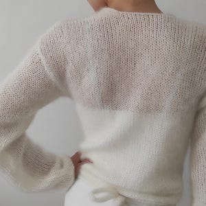 May include: A white, sheer knit sweater with long sleeves and a tie at the waist. The sweater is paired with a white skirt. A pearl hair clip is visible in the hair.