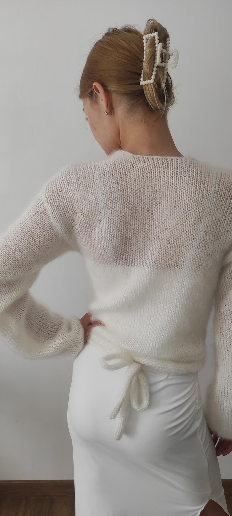 May include: A white, sheer knit sweater with long sleeves and a tie at the waist. The sweater is paired with a white skirt. A pearl hair clip is visible in the hair.
