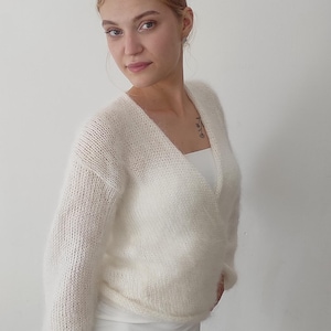 May include: A white, fluffy, wrap-style sweater with long sleeves. The sweater is paired with a white dress. The overall look is soft and feminine, suitable for layering or as a statement piece.