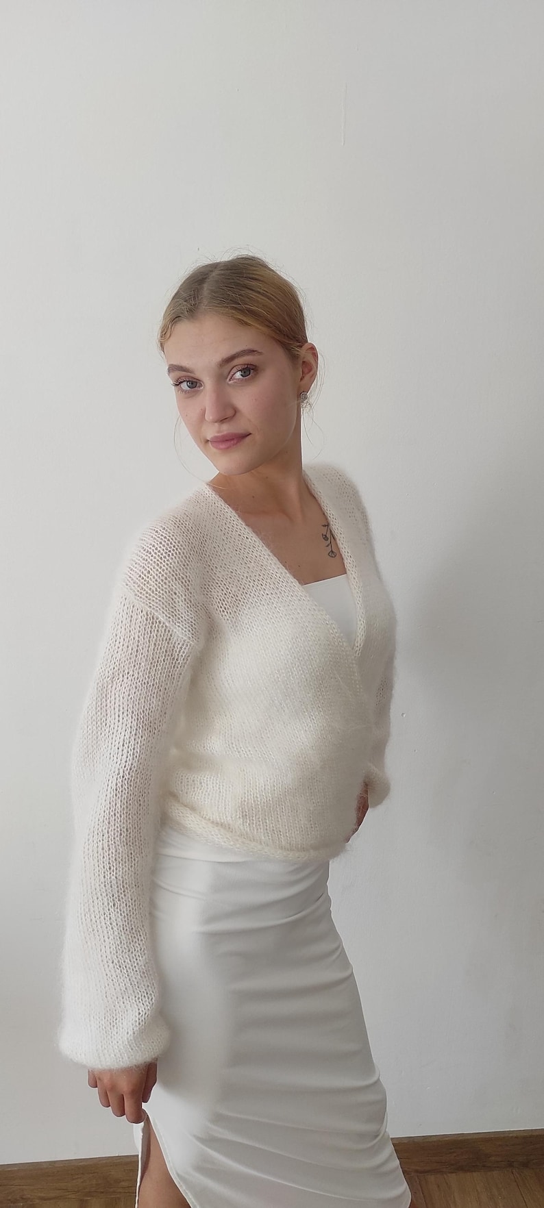 May include: A white, fluffy, wrap-style sweater with long sleeves. The sweater is paired with a white dress. The overall look is soft and feminine, suitable for layering or as a statement piece.