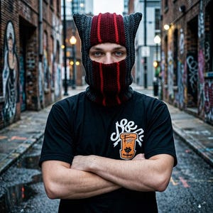 Könnte beinhalten: Eine gestrickte Sturmhaube in Grau, Burgunderrot und Rot, mit ohrenartigen Spitzen oben. Die Person trägt ein schwarzes T-Shirt mit einem weißen und orangefarbenen Aufdruck. Der Hintergrund ist eine Gasse mit Graffiti und Backsteingebäuden.