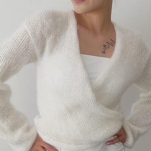 May include: A white, long-sleeved wrap sweater with a V-neck. The sweater is made of a soft, fuzzy material. The model is wearing a white top and skirt. The background is a plain white wall.