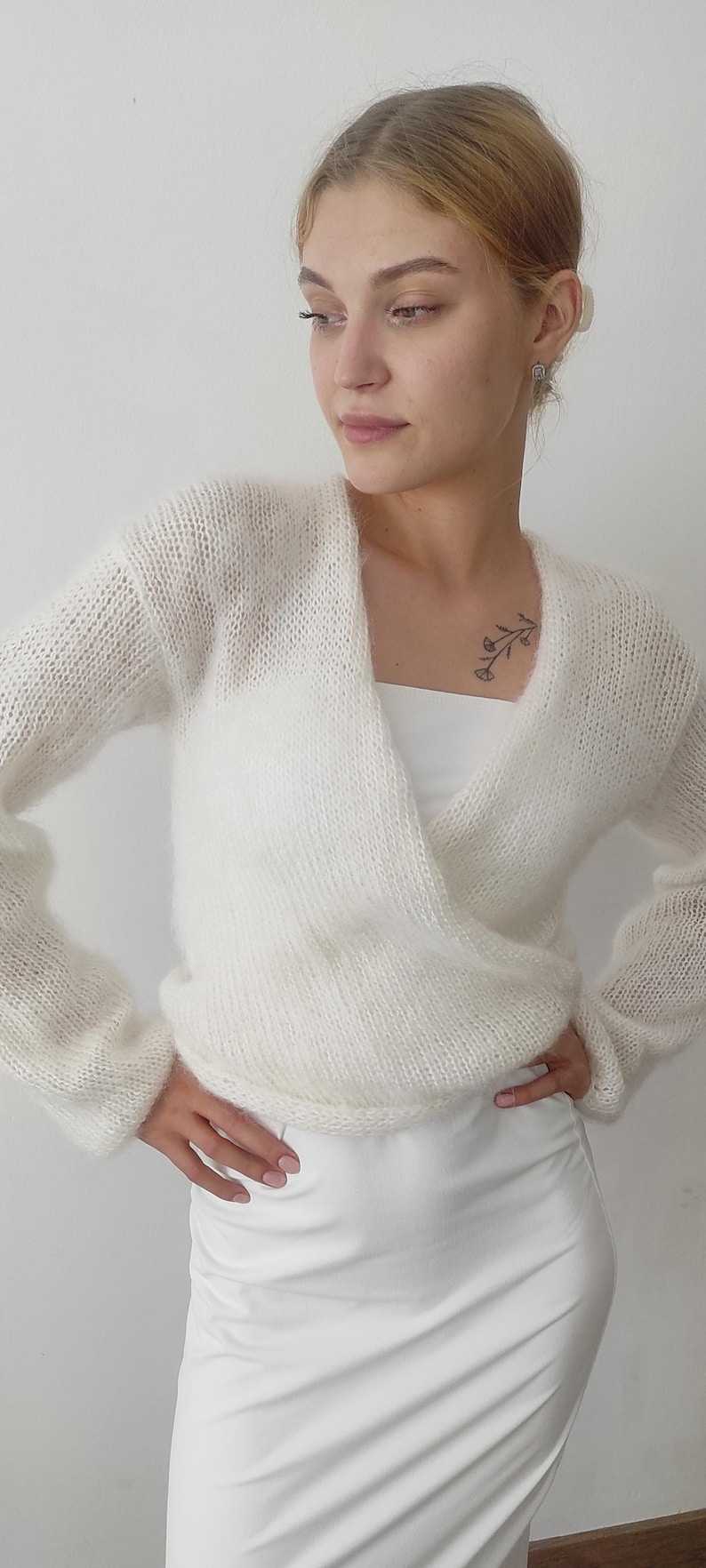 May include: A white, long-sleeved wrap sweater with a V-neck. The sweater is made of a soft, fuzzy material. The model is wearing a white top and skirt. The background is a plain white wall.