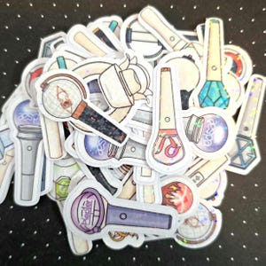 May include: A collection of holographic stickers featuring various designs of light sticks. The stickers showcase a range of colors, including silver, purple, blue, and pink. Some stickers have text, and the overall theme is related to K-pop.