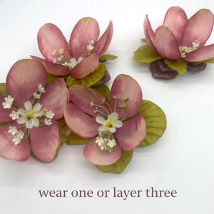 May include: Three artificial pink flower arrangements with green leaves and white accents. The flowers have a layered petal design and are arranged on a white surface. The text "wear one or layer three" is at the bottom.