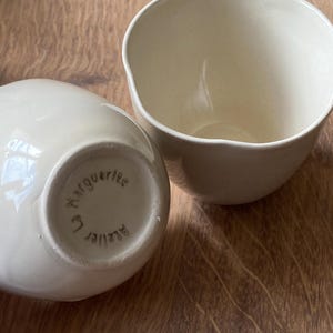 Peut inclure: Deux bols en céramique blanc cassé aux bords irréguliers sur une surface en bois. L'un est droit, l'autre est couché, révélant la base avec le texte "Le Marguerite Atelier" disposé en cercle.