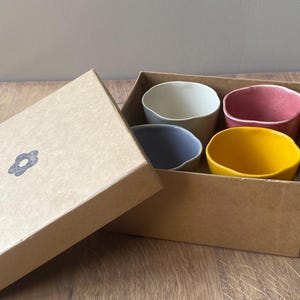 Peut inclure: Un ensemble de quatre petits bols en céramique colorés dans une boîte en carton. Les bols sont dans des tons de blanc, rose poudré, bleu-gris et jaune. La boîte est ouverte, révélant les bols à l'intérieur. La boîte a un motif en forme de fleur sur le couvercle.