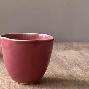 Peut inclure: Une petite tasse en céramique arrondie de couleur rose. La tasse a un bord légèrement irrégulier et une finition lisse et brillante. La tasse est posée sur une surface en bois.