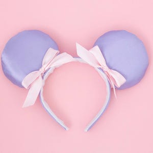 May include: A lavender-colored headband with two round ears and light pink bows. The ears are made of a satin-like material, and the headband is white and light blue. The background is a solid pink.