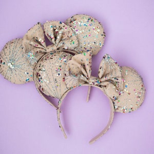 May include: A pair of beige mouse ears with a sparkly silver and multi-colored confetti design. The ears are attached to a headband.
